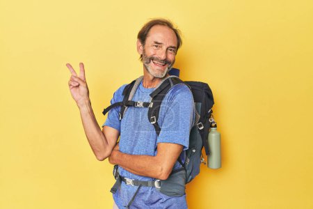 Photo for Middle-aged hiker with backpack in studio joyful and carefree showing a peace symbol with fingers. - Royalty Free Image