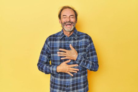 Téléchargez les photos : Caucasien d'âge moyen homme en studio jaune rit heureux et s'amuse à garder les mains sur l'estomac. - en image libre de droit