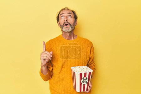 Téléchargez les photos : Homme jouissant de pop-corn en studio jaune pointant à l'envers avec bouche ouverte. - en image libre de droit