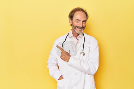 Téléchargez les photos : Docteur avec stéthoscope en studio jaune souriant et pointant du doigt, montrant quelque chose à blanc. - en image libre de droit