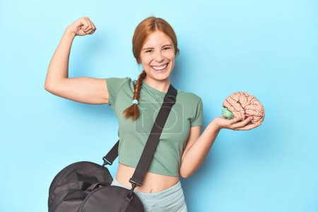 Photo for Sporty redhead with brain model on blue background raising fist after a victory, winner concept. - Royalty Free Image