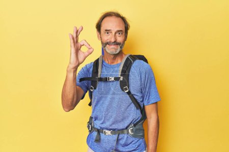 Photo for Middle-aged hiker with backpack in studio cheerful and confident showing ok gesture. - Royalty Free Image