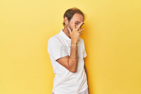 Foto de Caucásico hombre de mediana edad en el estudio amarillo parpadear en la cámara a través de los dedos, vergüenza cubriendo la cara. - Imagen libre de derechos