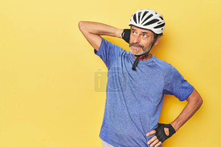 Foto de Ciclista con casco en estudio amarillo tocando la parte posterior de la cabeza, pensando y haciendo una elección. - Imagen libre de derechos