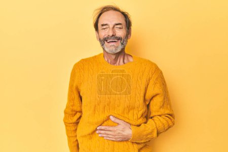 Téléchargez les photos : Homme d'âge moyen caucasien en studio jaune touche ventre, sourit doucement, manger et concept de satisfaction. - en image libre de droit