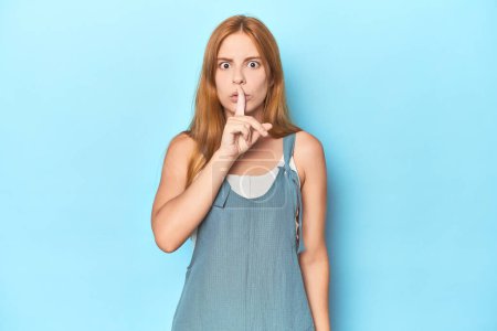 Téléchargez les photos : Rousse jeune femme sur fond bleu garder un secret ou demander le silence. - en image libre de droit