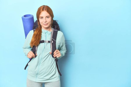 Photo for Mountaineer redhead with backpack and mat on blue background - Royalty Free Image