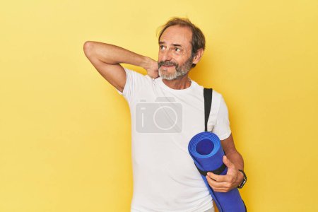 Téléchargez les photos : Homme avec tapis de yoga en studio jaune touchant l'arrière de la tête, pensant et faisant un choix. - en image libre de droit