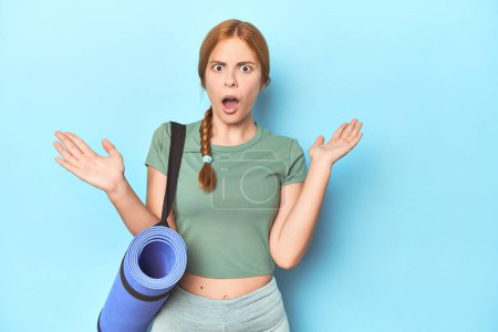 Photo for Redhead young woman holding yoga mat in studio surprised and shocked. - Royalty Free Image