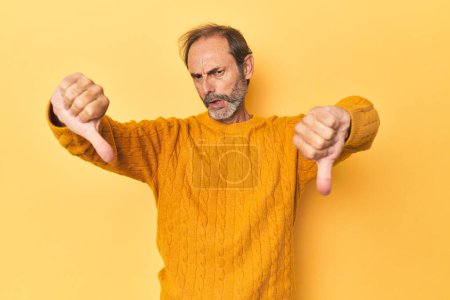 Foto de Caucásico hombre de mediana edad en estudio amarillo mostrando el pulgar hacia abajo y expresando aversión. - Imagen libre de derechos