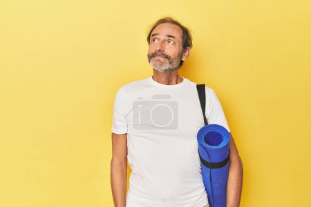 Téléchargez les photos : Homme avec tapis de yoga en studio jaune rêvant d'atteindre des objectifs et des buts - en image libre de droit