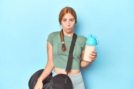 Photo for Redhead holding protein shake in blue studio shrugs shoulders and open eyes confused. - Royalty Free Image