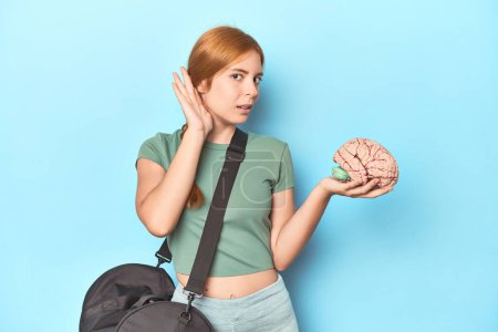 Photo for Sporty redhead with brain model on blue background trying to listening a gossip. - Royalty Free Image