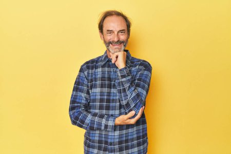Foto de Caucásico hombre de mediana edad en estudio amarillo sonriendo feliz y confiado, tocando la barbilla con la mano. - Imagen libre de derechos