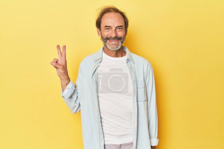 Foto de Caucásico hombre de mediana edad en estudio amarillo mostrando signo de victoria y sonriendo ampliamente. - Imagen libre de derechos