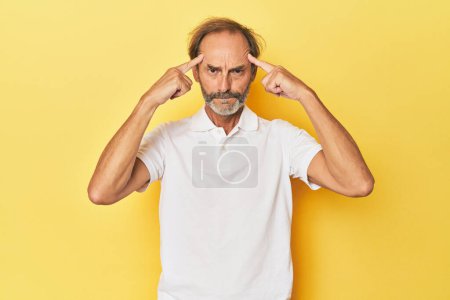 Téléchargez les photos : Homme d'âge moyen caucasien en studio jaune concentré sur une tâche, en gardant les index pointant la tête. - en image libre de droit