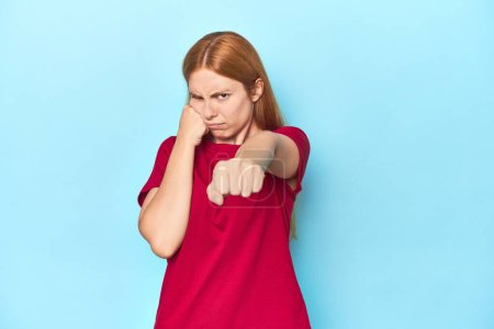 Téléchargez les photos : Jeune femme rousse sur fond bleu jetant un coup de poing, colère, combats à cause d'une dispute, boxe. - en image libre de droit