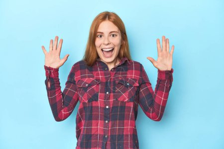 Photo for Redhead young woman on blue background receiving a pleasant surprise, excited and raising hands. - Royalty Free Image