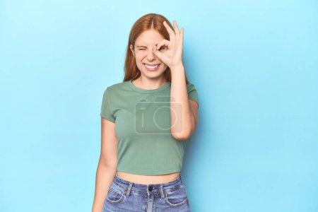 Téléchargez les photos : Rousse jeune femme sur fond bleu excité garder ok geste sur oeil. - en image libre de droit