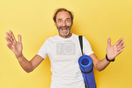 Photo for Man with yoga mat in yellow studio receiving a pleasant surprise, excited and raising hands. - Royalty Free Image