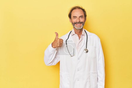 Foto de Doctor con estetoscopio en estudio amarillo sonriendo y levantando el pulgar hacia arriba - Imagen libre de derechos