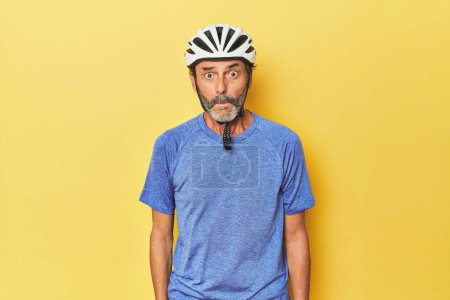 Foto de Ciclista con casco en estudio amarillo se encoge de hombros y los ojos abiertos confundido. - Imagen libre de derechos