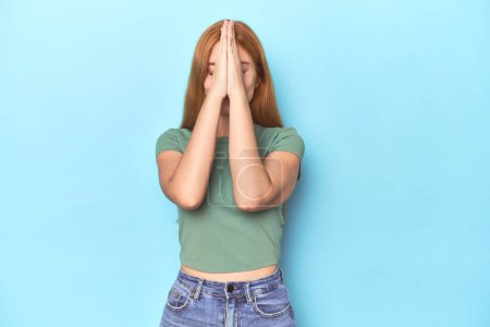 Téléchargez les photos : Rousse jeune femme sur fond bleu tenant la main dans la prière près de la bouche, se sent confiant. - en image libre de droit
