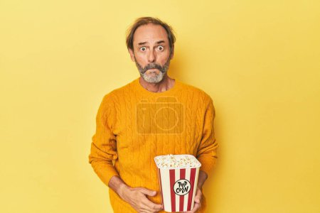 Foto de Hombre disfrutando de palomitas de maíz en estudio amarillo encoge hombros y los ojos abiertos confundido. - Imagen libre de derechos