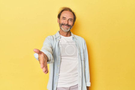 Téléchargez les photos : Blanc homme d'âge moyen en studio jaune étirant la main à la caméra dans le geste de salutation. - en image libre de droit