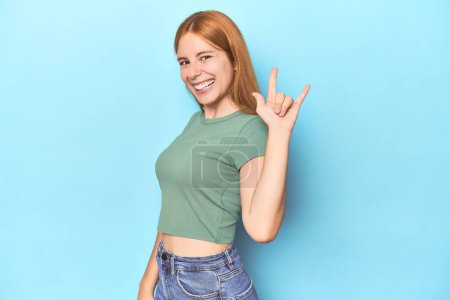 Photo for Redhead young woman on blue background showing a horns gesture as a revolution concept. - Royalty Free Image