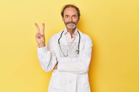 Foto de Doctor con estetoscopio en estudio amarillo mostrando el número dos con los dedos. - Imagen libre de derechos