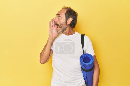 Foto de Hombre con esterilla de yoga en estudio amarillo gritando y sosteniendo la palma cerca de la boca abierta. - Imagen libre de derechos