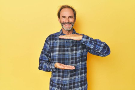 Foto de Caucásico hombre de mediana edad en estudio amarillo sosteniendo algo con ambas manos, presentación del producto. - Imagen libre de derechos