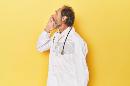 Foto de Médico con estetoscopio en estudio amarillo gritando y sosteniendo la palma cerca de la boca abierta. - Imagen libre de derechos