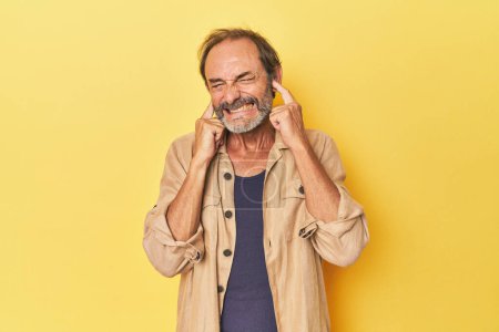 Foto de Caucásico hombre de mediana edad en estudio amarillo cubriendo orejas con las manos. - Imagen libre de derechos