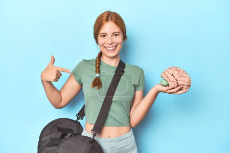 Photo for Sporty redhead with brain model on blue background person pointing by hand to a shirt copy space, proud and confident - Royalty Free Image