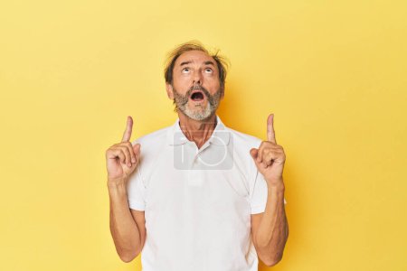 Téléchargez les photos : Blanc homme d'âge moyen en studio jaune pointant à l'envers avec bouche ouverte. - en image libre de droit