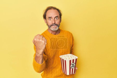 Photo for Man enjoying popcorn in yellow studio showing fist to camera, aggressive facial expression. - Royalty Free Image