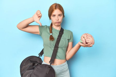 Photo for Sporty redhead with brain model on blue background showing a dislike gesture, thumbs down. Disagreement concept. - Royalty Free Image