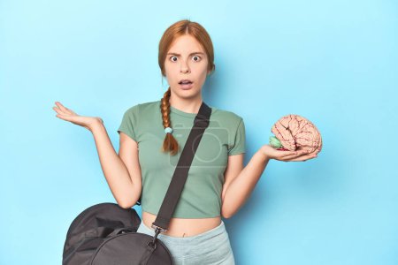 Photo for Sporty redhead with brain model on blue background surprised and shocked. - Royalty Free Image