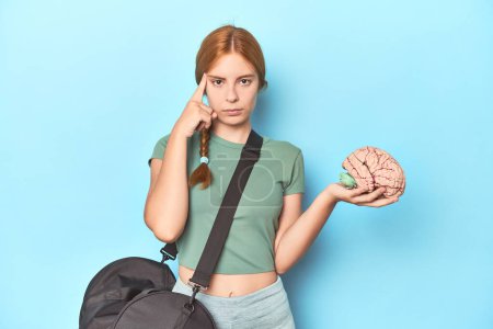 Photo for Sporty redhead with brain model on blue background pointing temple with finger, thinking, focused on a task. - Royalty Free Image