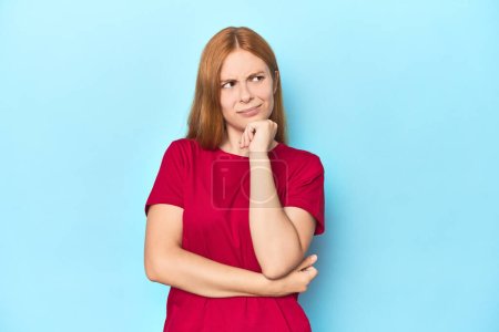 Photo for Redhead young woman on blue background thinking and looking up, being reflective, contemplating, having a fantasy. - Royalty Free Image