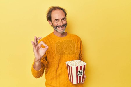 Photo for Man enjoying popcorn in yellow studio cheerful and confident showing ok gesture. - Royalty Free Image