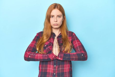 Foto de Pelirroja joven sobre fondo azul rezando, mostrando devoción, persona religiosa buscando inspiración divina. - Imagen libre de derechos