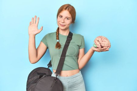 Photo for Sporty redhead with brain model on blue background smiling cheerful showing number five with fingers. - Royalty Free Image