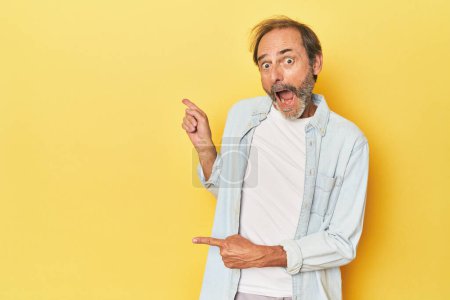 Téléchargez les photos : Homme d'âge moyen caucasien dans un studio jaune pointant avec les index vers un espace de copie, exprimant l'excitation et le désir. - en image libre de droit