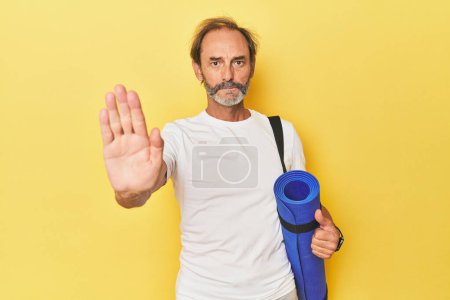 Foto de Hombre con esterilla de yoga en estudio amarillo de pie con la mano extendida mostrando señal de stop, lo que le impide. - Imagen libre de derechos