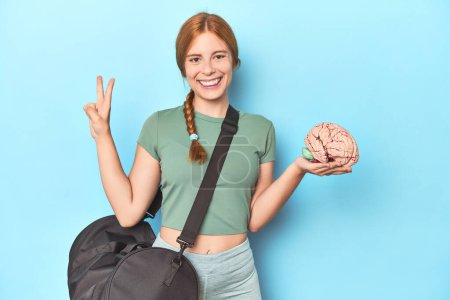 Photo for Sporty redhead with brain model on blue background showing number two with fingers. - Royalty Free Image