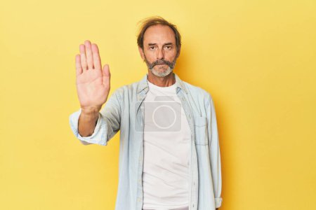 Téléchargez les photos : Homme d'âge moyen caucasien en studio jaune debout avec la main tendue montrant le panneau stop, vous empêchant. - en image libre de droit
