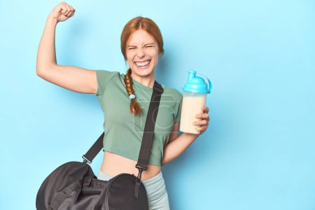 Téléchargez les photos : Redhead tenant shake protéine en studio bleu levant le poing après une victoire, concept gagnant. - en image libre de droit